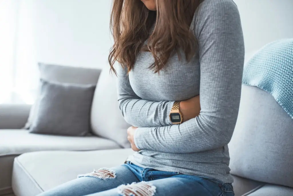 A woman sitting on a couch with her arms folded across her stomach, looking unwell