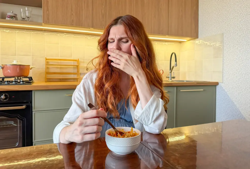 A woman sitting at a kitchen table covering her mouth, unable to face the bowl of food in front of her