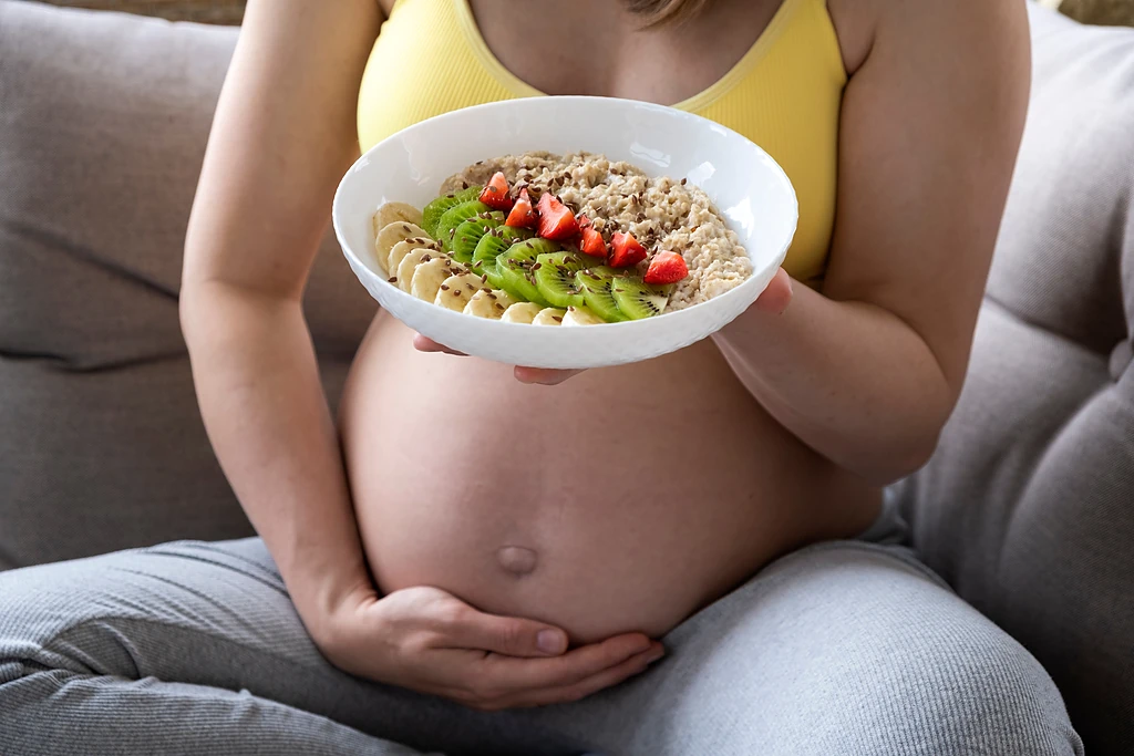 A pregnant woman sitting on a couch holding a bowl of porridge topped with banana, kiwifruit and strawberries